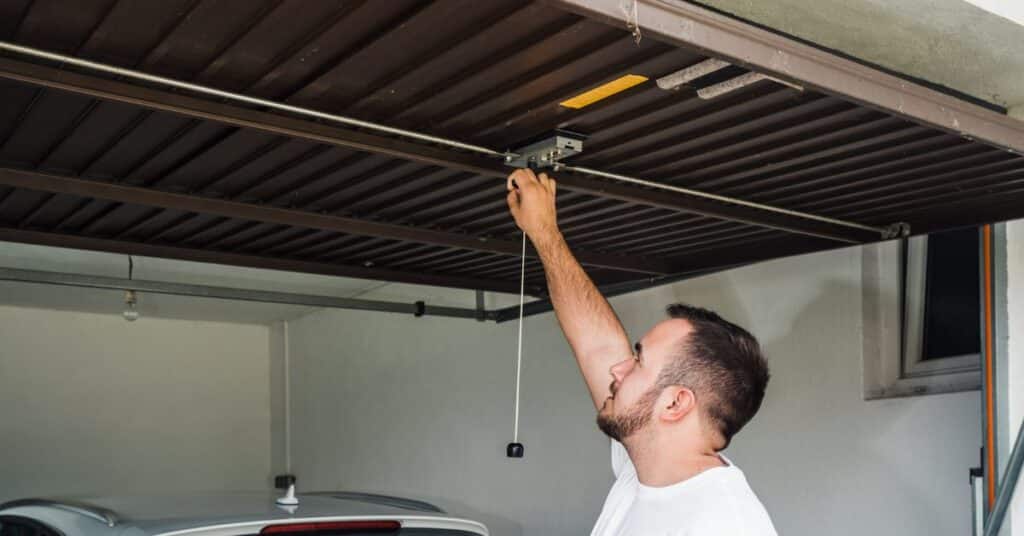A man in a white shirt reaches up to manually open a brown garage door using the emergency release cord.